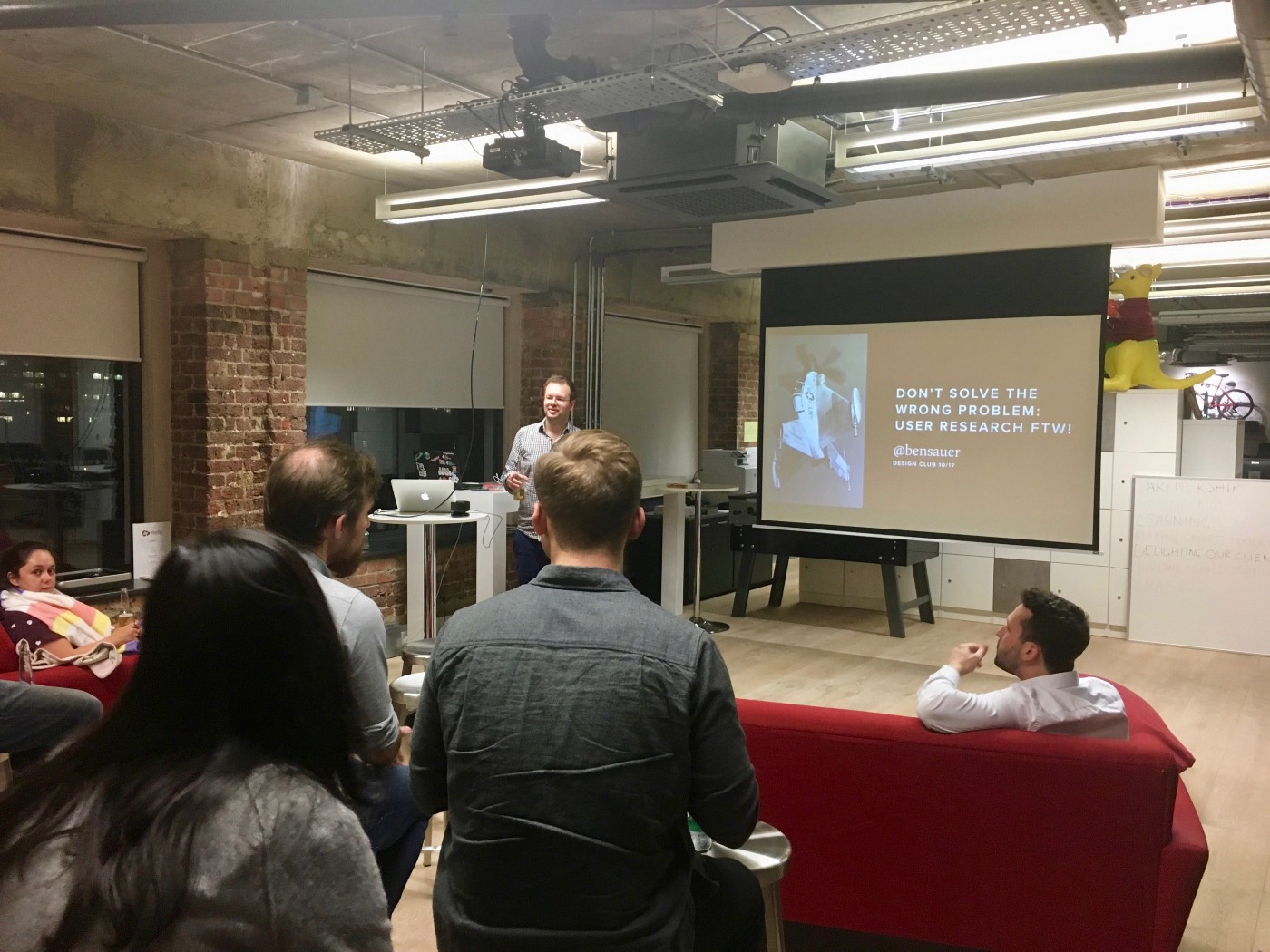 Ben addresses some people, behind him his slides projected onto a large monitor