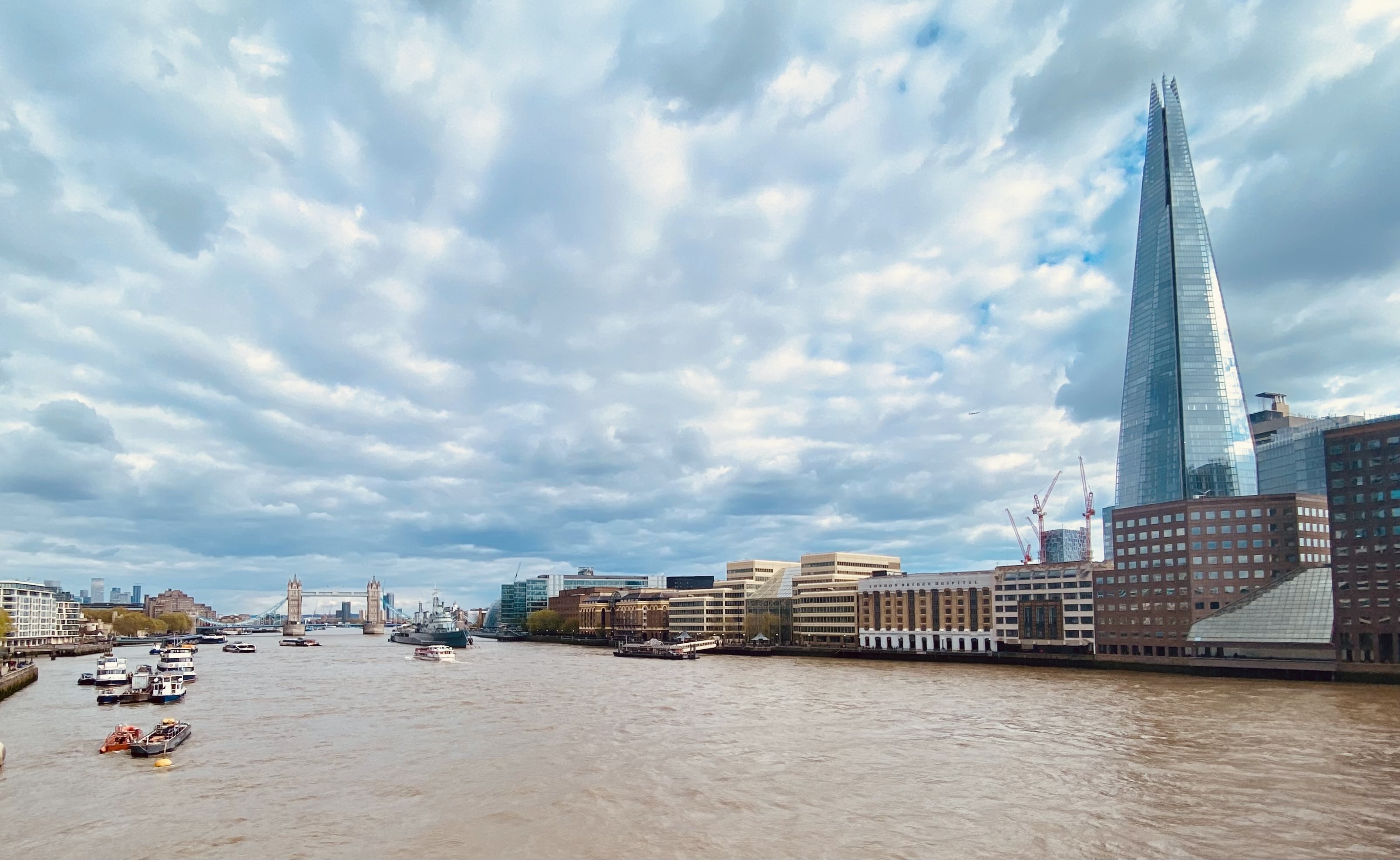The view from London Bridge. A huge cloudy sky stretches out over the river below. The Shard can be seen on the right and Tower Bridge in the distance.