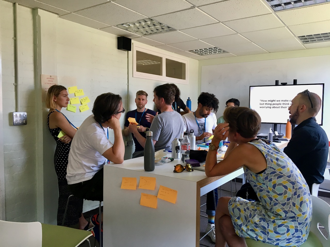 People gathered in a room, some sat around a table some standing with post-its strewn around the walls